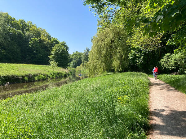 Auf dem Travewanderweg sind Radfahrer unterwegs, auf der Trave fahren Kajakfahrer